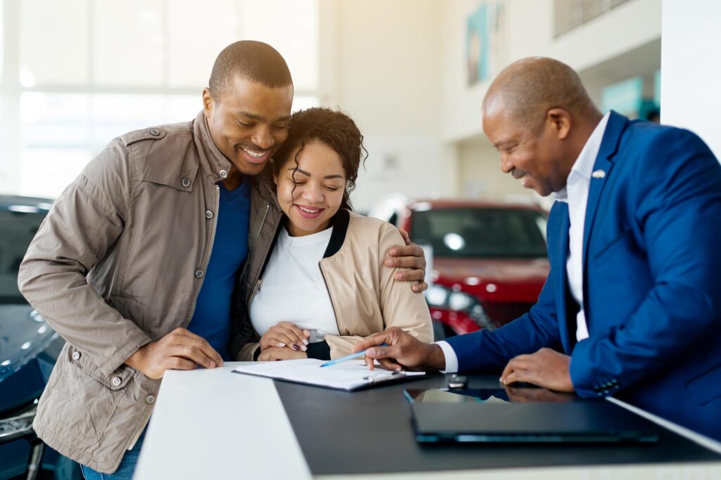 A young couple smiles as they review and sign a car purchase agreement at a dealership. The salesman provides guidance and support during the transaction.
