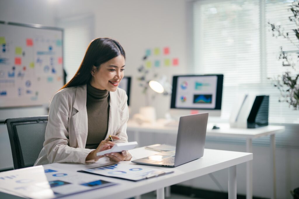 Smiling businesswoman calculating and analyzing financial data on a laptop, engaging with graphs and charts while working in a modern office environment, showcasing professionalism and expertise. She's content knowing she is working with the right payment partner.