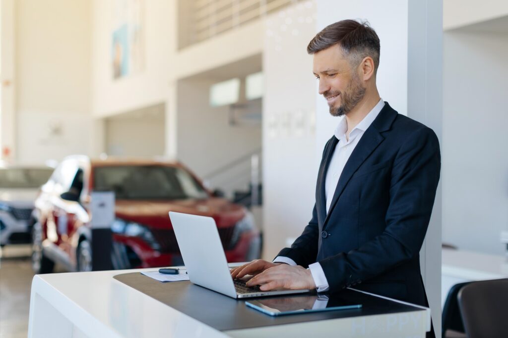 Positive car salesman using virtual terminal on his laptop computer at work desk in auto dealership center. Cheerful millennial automobile manager working with pc in showroom store, free space